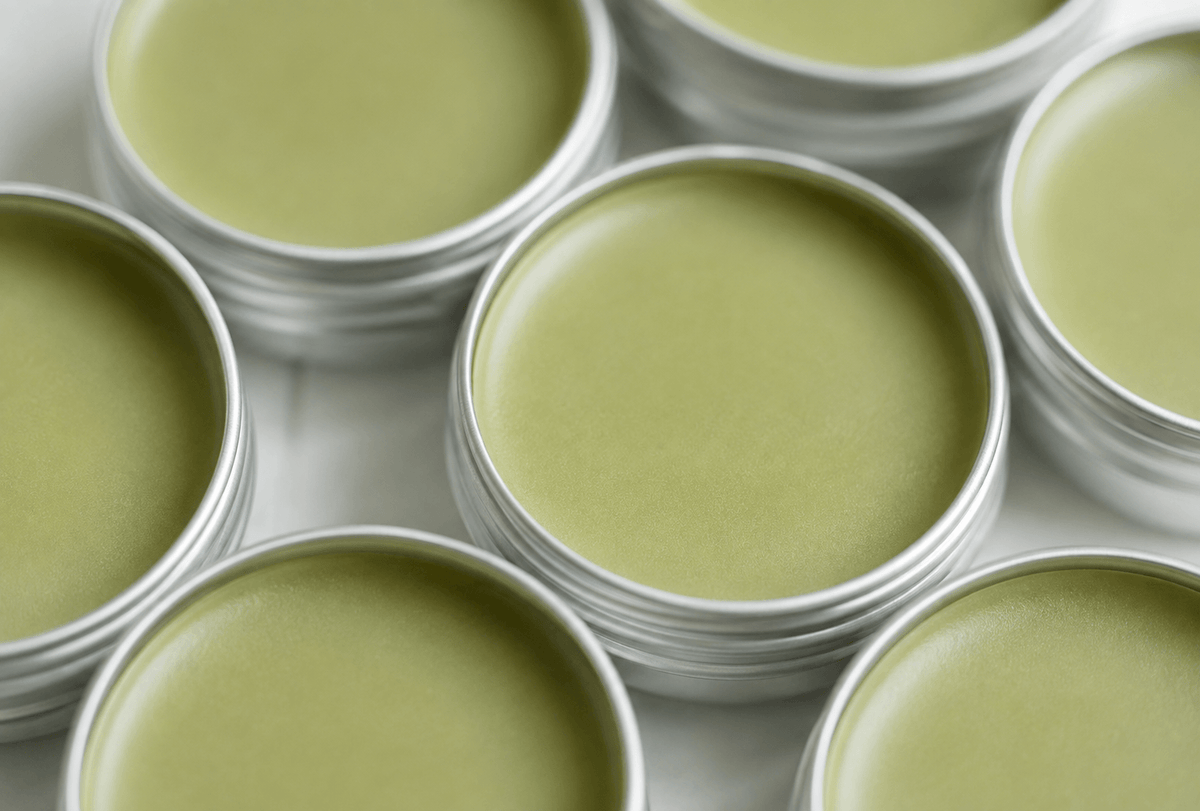 Close-up of several round silver metal tins filled with a pale matcha-green balm or cream, arranged on a light surface.