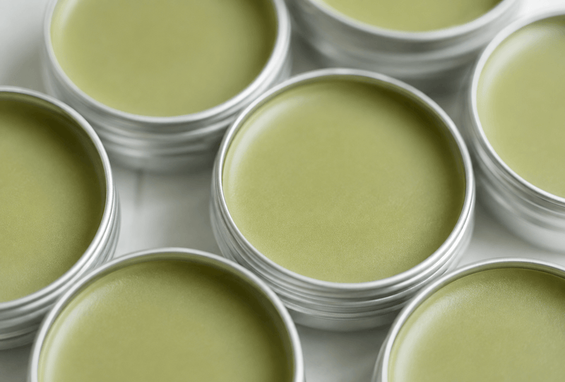 Close-up of several round silver metal tins filled with a pale matcha-green balm or cream, arranged on a light surface.