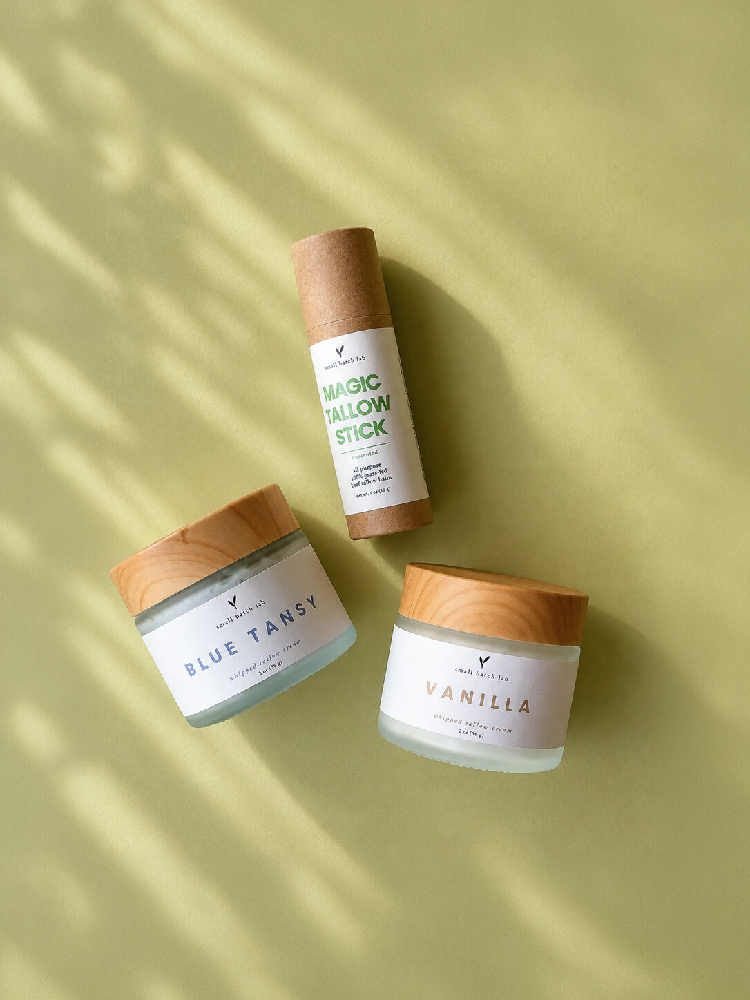 Three skincare products on a light green surface. A tube labeled "Magic Tallow Stick" and jars with "Blue Tansy" and "Vanilla" text, all with natural wood lids.
