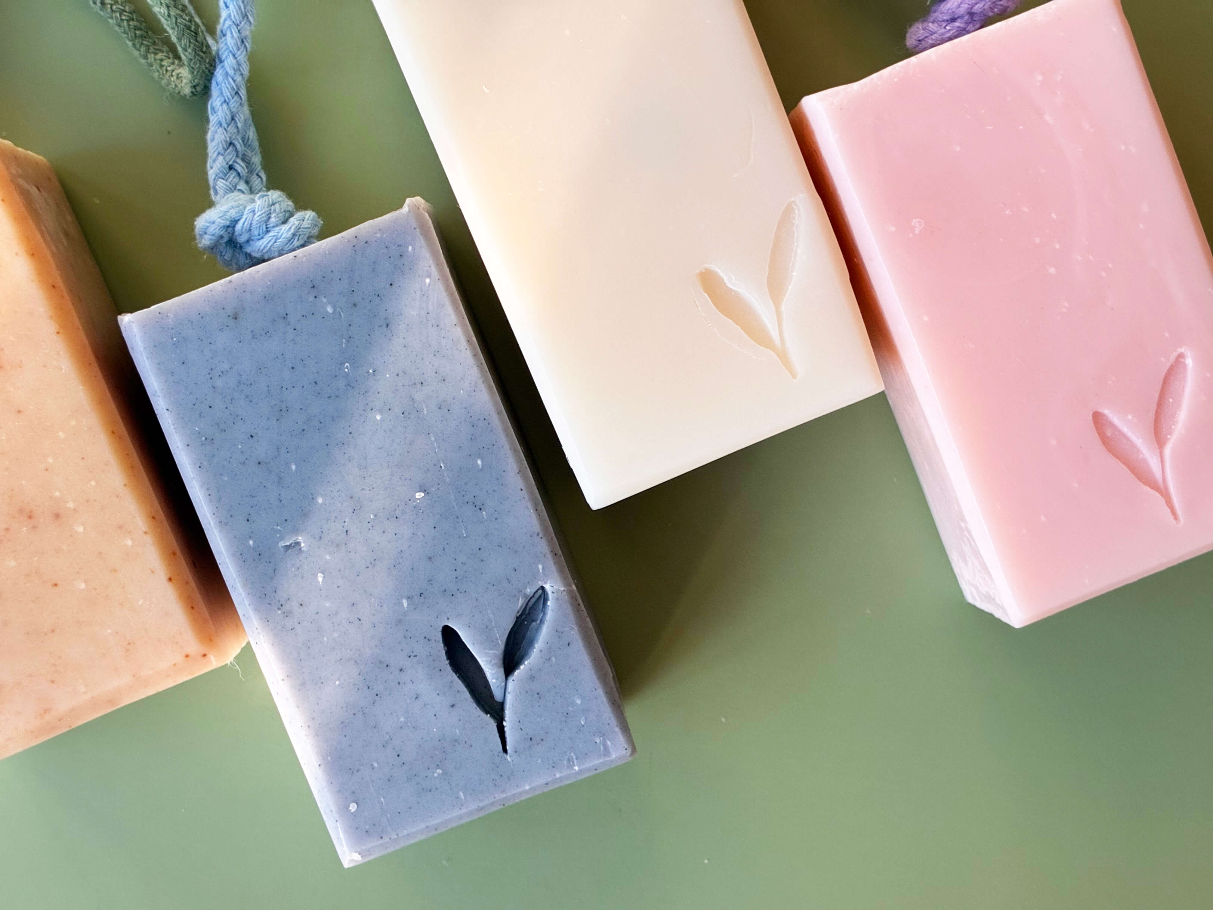 Four rectangular handmade soap bars with leaf-shaped indentations, arranged diagonally on a green surface. The soaps are in peach, dusty blue, cream, and light pink, each with a small hanging loop at the top.