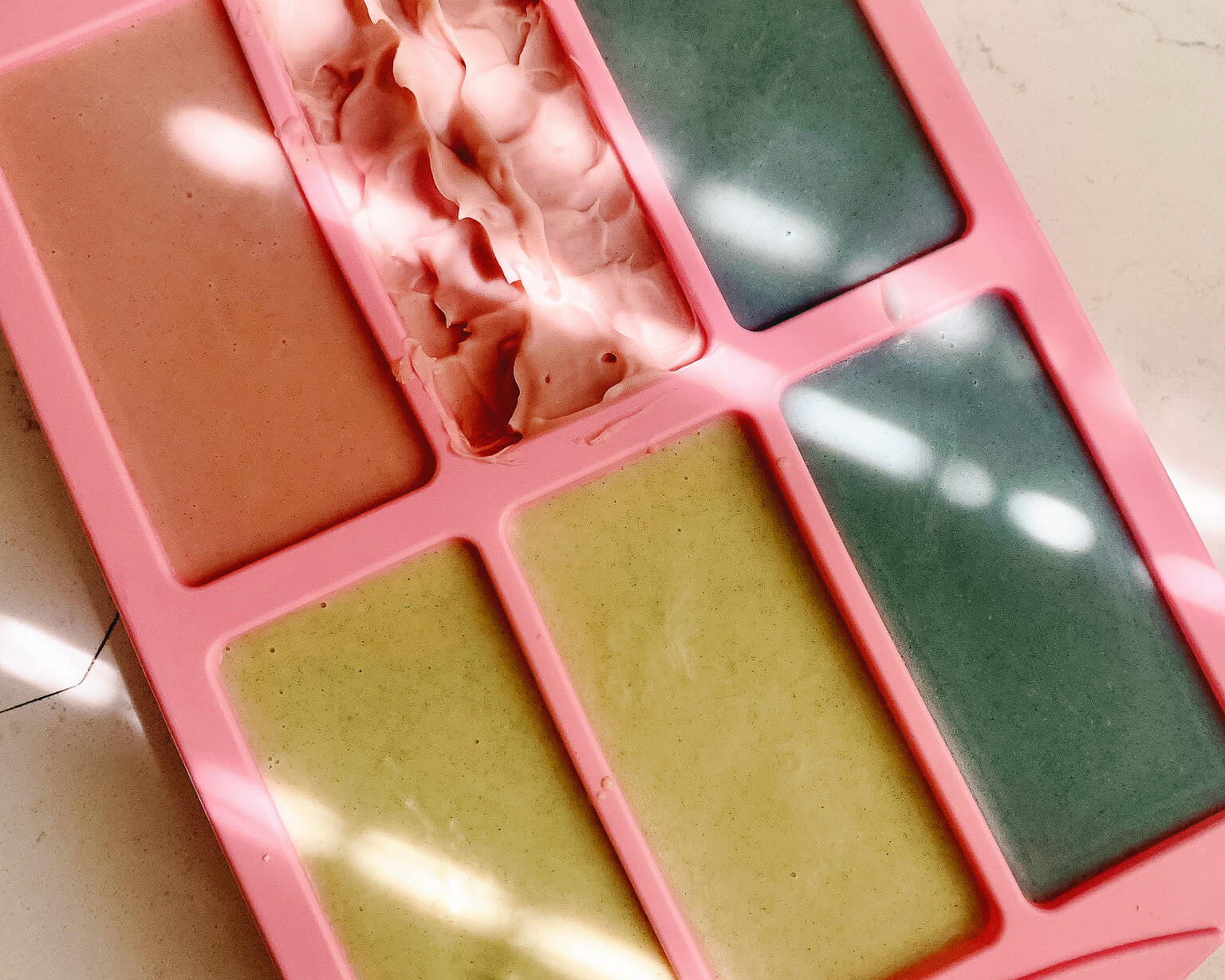 Pink soap mold with various colored soap bars inside on a light surface.